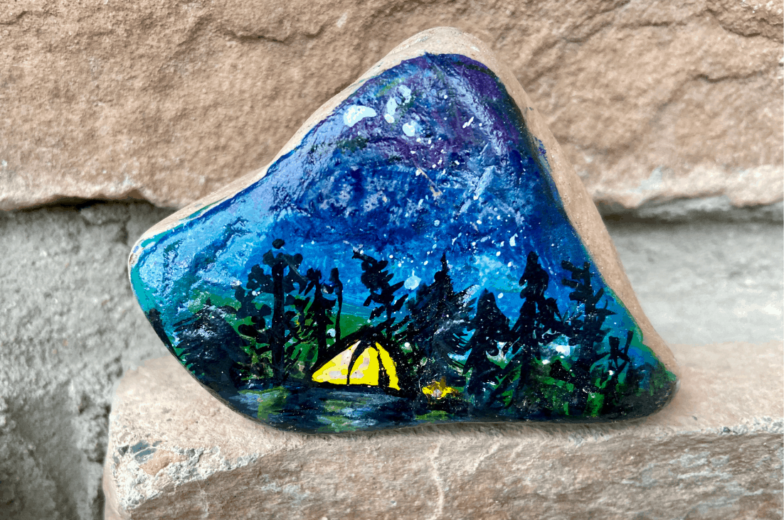 A rock painted with a beautiful night time camping scene with a lit tent and a little campfire