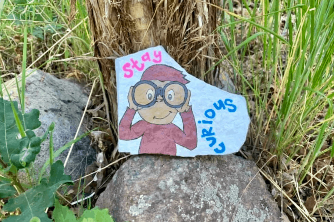 A Curious George painted rock. It says Stay Curious 